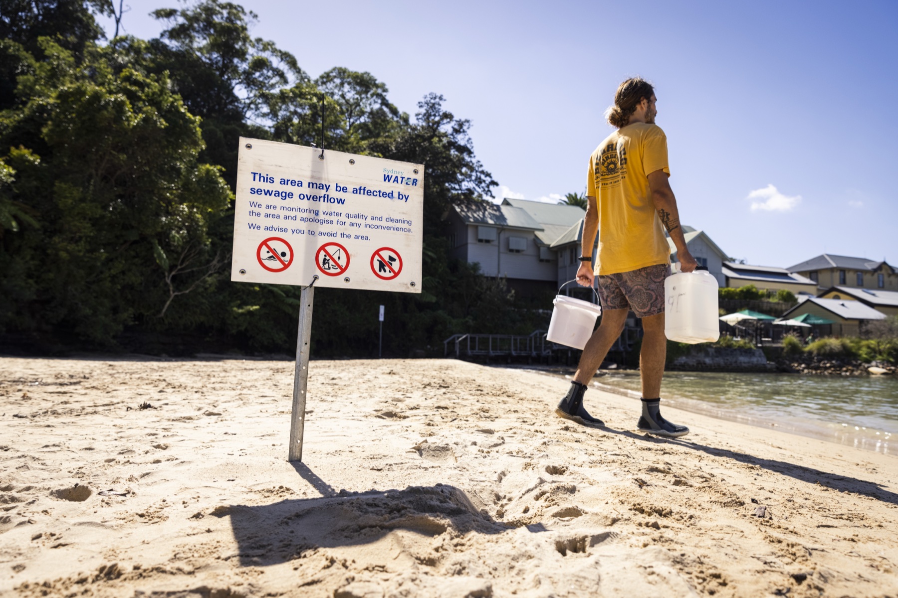 Carrying sampling containers beside a coastal sewage-overflow warning sign